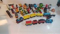 Photo showing multiple wooden and metal Thomas and Friends trains and cars arranged on a white surface, displaying different colors and characters.