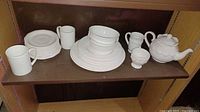 Shelf with white dinnerware including plates, bowls, cups, teapot, creamer, and sugar bowl.