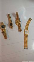 Four gold tone women's wrist watches laid out on a white surface, showing metal link bands and various dial sizes and colors.