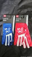 Two pairs of new Under Armour UA Heater batting gloves, one blue and one red, shown with packaging tags.