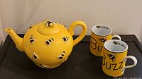Yellow honey bee-themed ceramic teapot with two matching mugs on black surface.