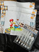 Four cloth placemats and packaged cutlery set displayed together on dark fabric, showing Disney characters and plaid borders.