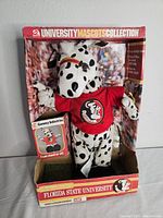 Full view of the Florida State University mascot plush in original display box showing the plush wearing a red shirt with the FSU logo and spots all over the body, set against a stadium crowd background.