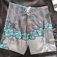 Hard Rock boardshorts shown from front angle with visible front lace-up closure, floral and guitar print, side cargo pocket with logo patch, and tags attached.