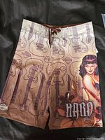 Photo showing front side of Hard Rock boardshorts with pinup girl graphic, guitars, and 'HARD' wording in shades of brown and beige.