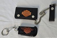 Photo of a black leather Harley Davidson change wallet with snap button closure and attached metal chain, alongside a black leather lighter case with whip stitching and a Harley Davidson logo.