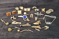 Top-down view of assorted cufflinks and tie clips laid out on black fabric, showing variety of shapes, colors, and designs