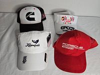 Four assorted hats displayed on white background; Cummins black and white trucker hat, white Kamasali baseball cap with tags, white EPCOT visor, red Porsche Motorsport hat.