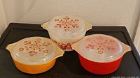 Three vintage Pyrex bowls with handles and matching lids, orange, red and white colors, floral and bird pattern.