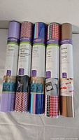 Photo showing five rolls of Cricut brand iron-on vinyl rolls including lavender, black and red pattern, and rose gold metallic.