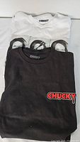 Photo showing folded white and black Chucky T-shirts stacked, highlighting the brand name inside the collars.