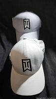 Front view of two white Tiger Woods golf hats stacked, showing black and white TW logos
