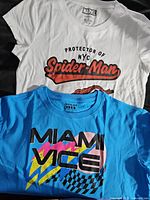 Photo showing both t-shirts laid flat: white Spider-Man t-shirt on top and blue Miami Vice t-shirt underneath.