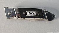 Side view of closed folding knife showing SOG logo on black G10 textured handle and stainless steel bolsters.
