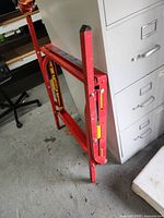 Red folding metal table saw stand by Bosch, folded and standing upright beside a filing cabinet.