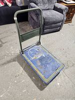 Metal collapsible push cart with a green frame and blue platform. Shows overall wear and usage.