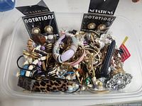 Photo showing a large clear plastic container filled with various costume jewelry items including bracelets, rings, earrings, and brooches in multiple colors and materials.
