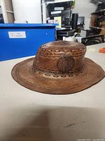 Side view of the brown leather hat showing embossed design and wide brim, placed on a white table with a blue bin in the background.