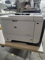 Front and side view of the HP LaserJet P3015 printer on a table, showing power indicator lit.