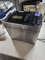 Front side view of the stainless steel KBS bread maker on a table, showing the overall appliance including handle and part of the power cord.