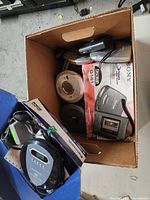 Top-down photo of a cardboard box containing multiple portable CD and tape players, including a boxed Sony tape player and loose CD players in various colors and brands.