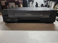 Front view of Toshiba VHS VCR showing tape loading slot, buttons, and branding.