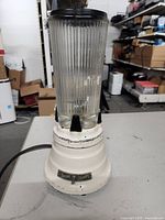 Front view of vintage Waring blender showing ribbed glass jar with black lid and metal base with some paint wear.