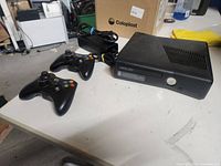 Xbox 360 Elite console with two black wireless controllers and power cable on a white table.