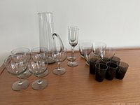 Photo showing a collection of various Danish Holmegaard glassware including a tall pitcher, multiple clear and smoky gray glasses arranged on a wooden surface.