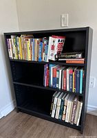Dark wood coloured IKEA bookcase filled with books on three shelves, showing adjustable shelf holes on inner sides.