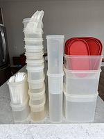 Photo of stackable translucent plastic food storage containers in various sizes, some with red lids and white measuring spoons on top, arranged neatly on a countertop.