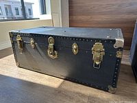 Photo of a black rectangular steamer chest with brass corner protectors and locks, placed on wooden floor with natural light.