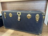 Photo of a black English steamer chest with brass accents, showing front exterior with locked latch and hardware details.