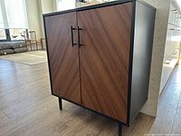 Photo of storage cabinet showing walnut finish doors, black metal handles, black metal legs and side panels.
