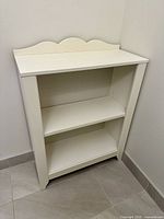 White painted wooden bookcase placed in corner against wall, showing two shelves and scalloped decorative back panel.