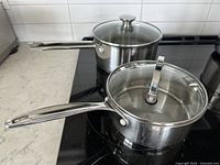 Two stainless steel pans placed on a stovetop, each having a glass lid with a knob on top and long stainless steel handles.