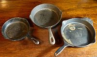 Three cast iron pans on a wooden surface including the Wagner Ware skillet on the left, a smaller skillet in the center, and a pan with side handles on the right.