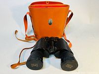 Binoculars and their brown leather carrying case with shoulder strap. Case has silver clasp and orange-red interior lining.