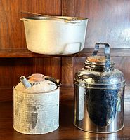 Photo showing large metal cook pot on a shelf behind two metal buckets and a shiny metal canister with wooden lid and handle.