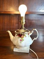 Overall front view of the Royal Albert 'Old Country Roses' teapot lamp, showing floral ceramic teapot on marble base with lit bulb on top.