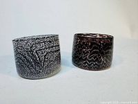 Two handblown glass tumblers, one predominantly white with black abstract pattern and the other darker with white pattern, shown from side angles.
