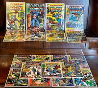 Overall view of 17 Captain America comic books laid out on a wooden surface, five upright in protectors and twelve flat.