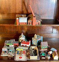 Overview photo of the collection of HO scale vintage buildings and accessories arranged on a wooden shelf, showing a variety of structures including commercial buildings, houses, a church, vehicles, and scenic elements.