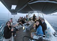 Group of people sitting and enjoying on a covered deck of a sailboat on water with city skyline in background.