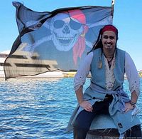 Person dressed as pirate sitting in front of pirate flag skull and crossbones at waterfront.