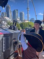 Photo of child steering the helm on a pirate ship with an adult in costume, Toronto city skyline and CN Tower visible.