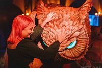 Photo of attendee interacting with large fantasy owl creature prop with glowing blue eyes under warm lighting.