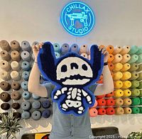 Person holding a blue outline skeleton tufted rug in front of a colorful yarn wall at Chillax Studio.
