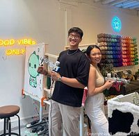 Two people holding tufting guns standing side by side in front of a tufting frame with a cartoon character design, neon sign stating 'GOOD VIBES ONLY' and yarn spools on a wall rack.