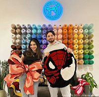 Two adults holding handmade tufted rugs shaped like a big red bow and a Spider-Man face, standing in front of a wall with multiple rows of colorful yarn skeins and a neon Chillax Studio sign.